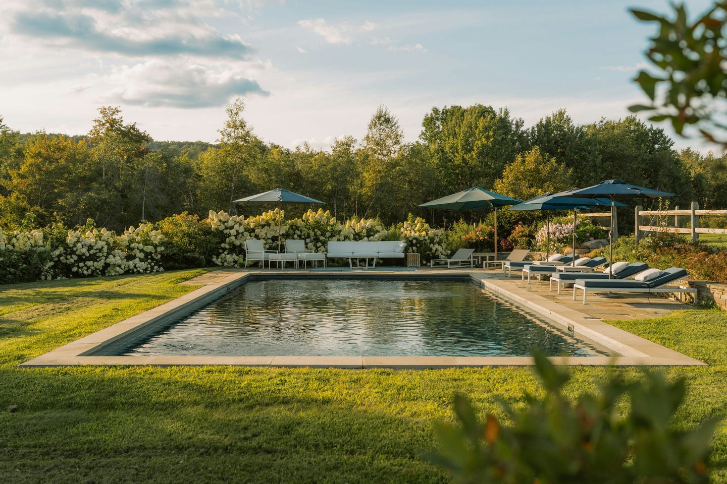 A long lawn and stone-edged pool with hydrangeas and loungers in evening light